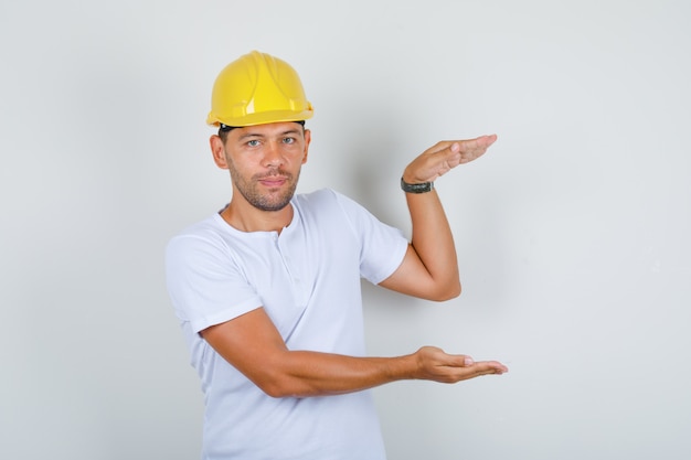 Builder man doing large size sign with hands in white t-shirt, helmet, front view.