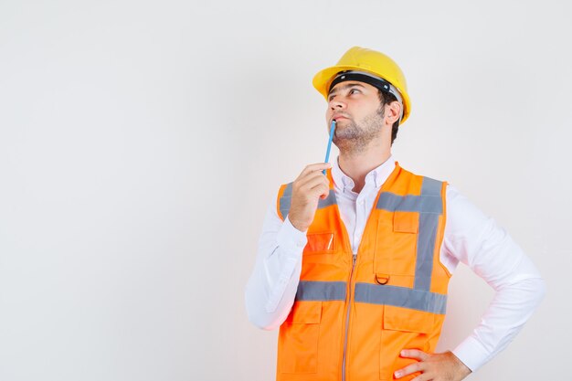 Builder man holding pencil near mouth in shirt, uniform and looking pensive. front view.