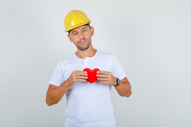 Строитель мужчина держит красное сердце в белой футболке, шлеме и выглядит счастливым, вид спереди