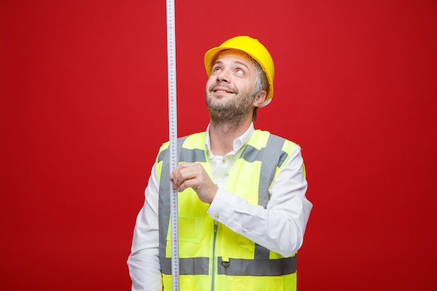 赤い背景の上に立っている幸せそうな顔に笑顔で見上げる測定テープを保持している建設制服と安全ヘルメットのビルダーの男