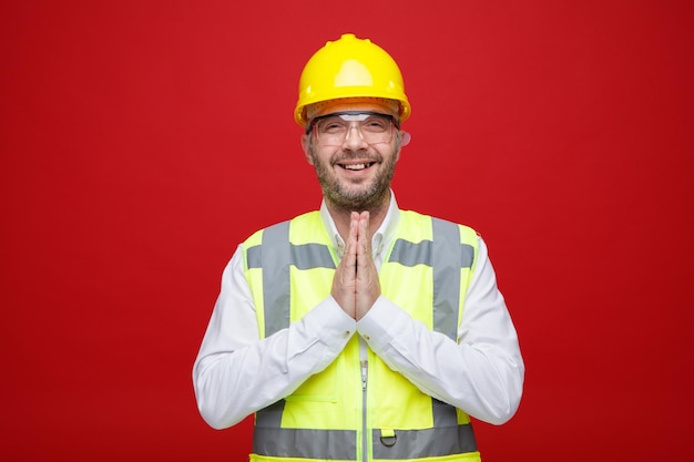 건설 유니폼과 안전 헬멧을 쓴 건축업자가 분홍색 배경 위에 손바닥을 함께 들고 즐겁게 웃고 있는 카메라를 바라보고 있습니다.