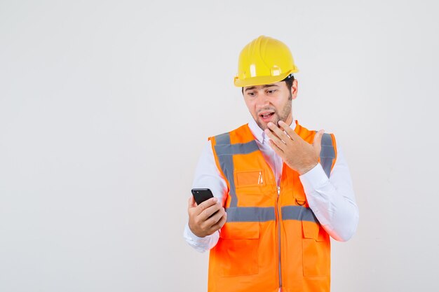 Builder man looking at smartphone with hand on chin in shirt, uniform and looking surprised. front view.