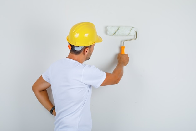 Builder man painting wall with roller in white t-shirt, helmet and looking busy, back view.
