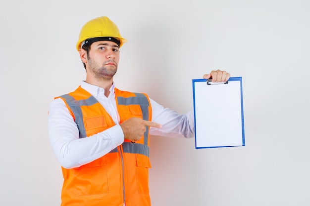 Builder man pointing finger at clipboard in shirt, uniform and looking serious. front view.