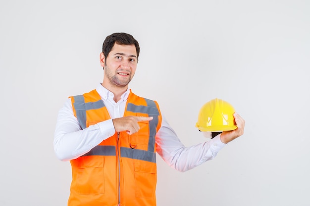 Builder man pointing finger at helmet in shirt, uniform and looking cheery , front view.