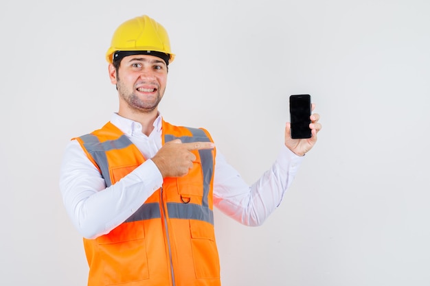 Builder man pointing finger at smartphone in shirt, uniform and looking cheery , front view.
