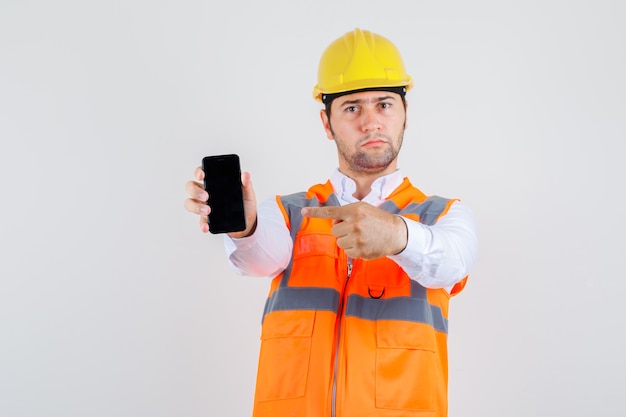 Builder man pointing finger at smartphone in shirt, uniform and looking serious , front view.