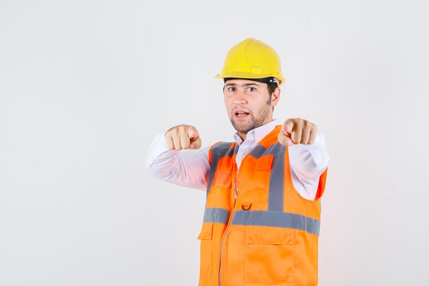 Builder man pointing fingers in shirt, uniform and looking confident. front view.