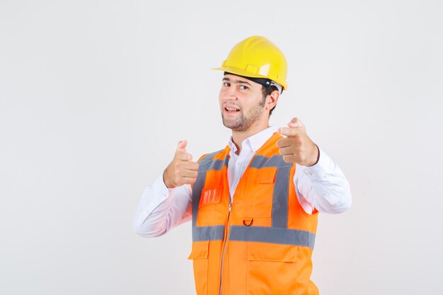 Builder man pointing to invite in shirt, uniform and looking positive. front view.