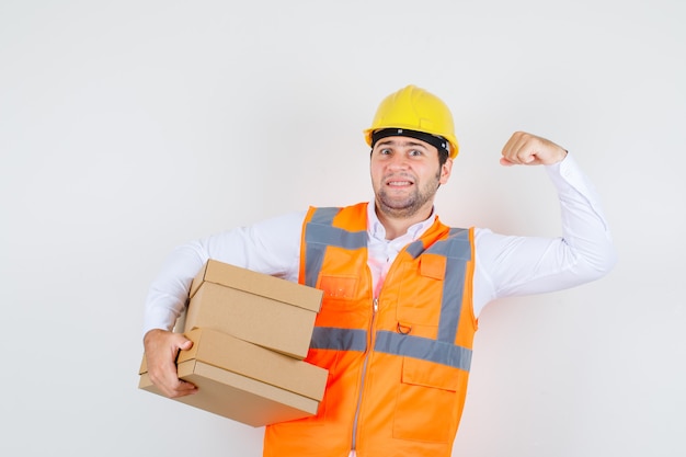 Builder man in shirt, uniform holding cardboard boxes while showing muscle and looking confident , front view.