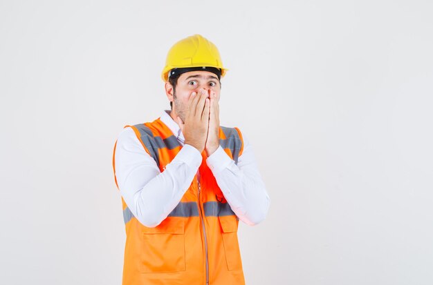 Builder man in shirt, uniform holding hands on mouth and looking scared , front view.