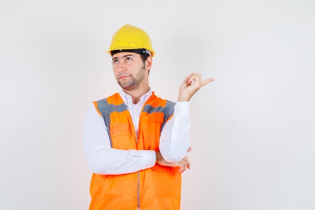 Builder man in shirt, uniform looking up while pointing away and looking pensive , front view.