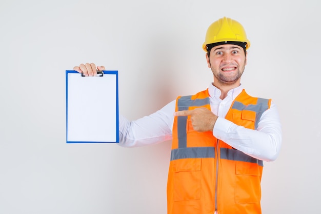 Builder man in shirt, uniform pointing finger at clipboard and looking joyful , front view.