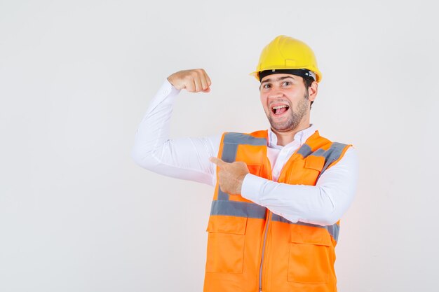 Builder man in shirt, uniform pointing at his muscle and looking strong , front view.