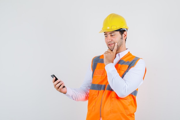 Builder man in shirt, uniform thinking while looking at smartphone and looking positive , front view.