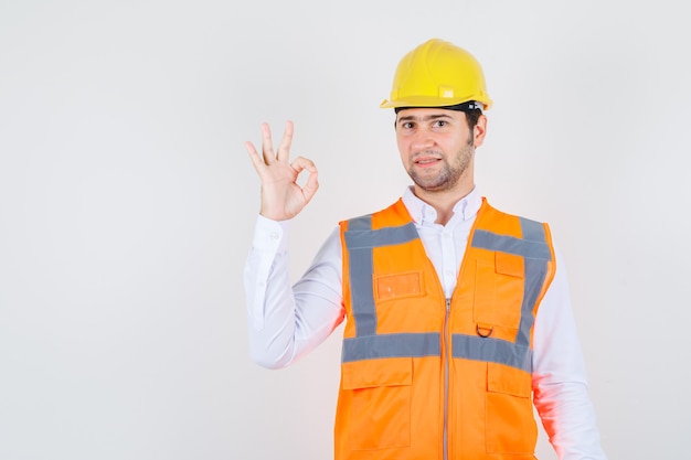 Builder man showing ok gesture in shirt, uniform and looking glad , front view.