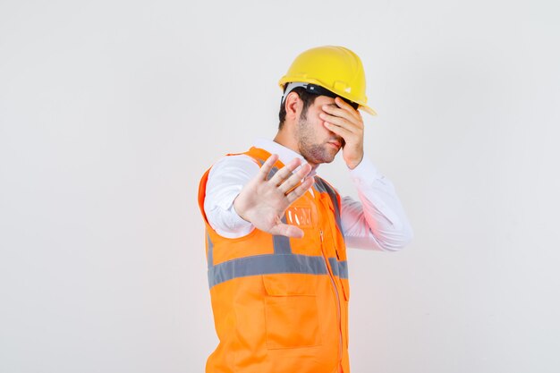 Builder man showing stop gesture while covering eyes in shirt, uniform front view.