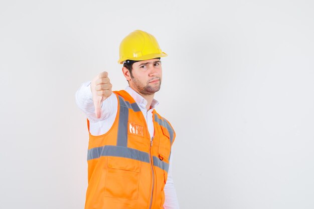 Builder man showing thumb down in shirt, uniform and looking displeased. front view.