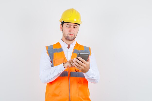 Builder man using calculator in shirt, uniform and looking busy , front view.