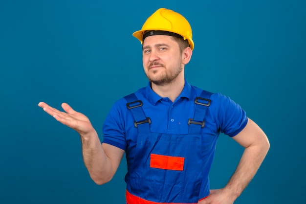 Бесплатное фото Человек-строитель в строительной форме и защитном шлеме, указывая в сторону с открытой ладонью руки, демонстрирует пространство для копирования, уверенно улыбаясь над изолированной синей стеной