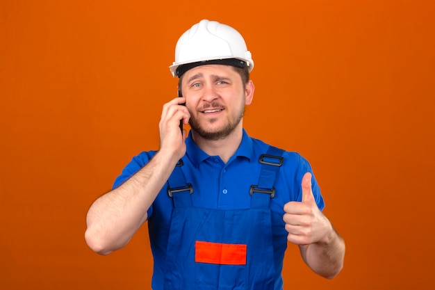 Бесплатное фото Человек-строитель в строительной форме и шлеме безопасности разговаривает по мобильному телефону, улыбаясь, показывая большой палец на камеру, стоящую над изолированной оранжевой стеной