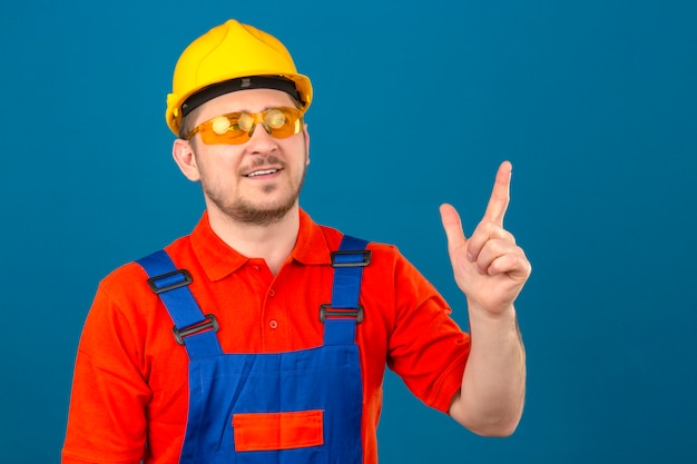 Builder man wearing construction uniform glasses and security helmet looking up pointing with finger having new idea standing over isolated blue wall