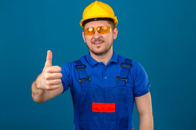 Foto gratuita pollice di mostra amichevole sorridente d'uso del casco di sicurezza dell'uomo d'uso del casco del costruttore e di sicurezza su che controlla parete blu isolata