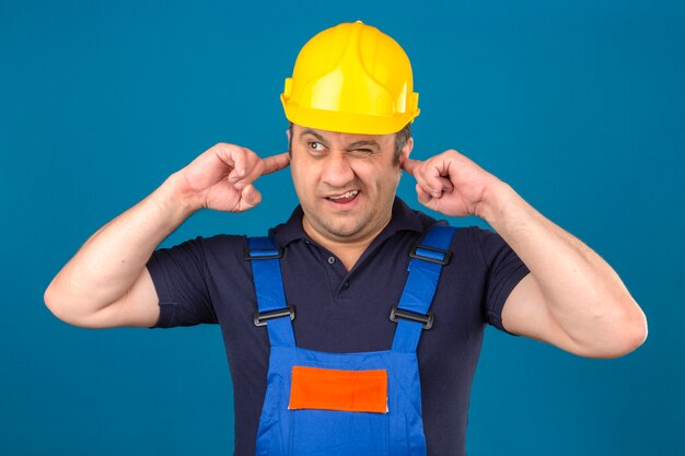 孤立した青い壁にノイズが原因で建設の制服と安全ヘルメットを閉じる耳を身に着けているビルダー男