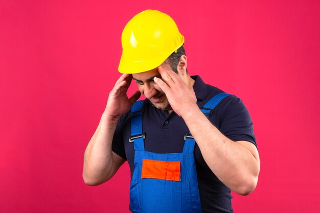 Builder man wearing construction uniform and safety helmet looking tired and overworked touching head feeling headache standing over isolated pink wall