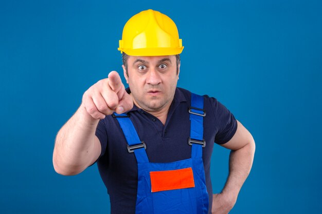 ビルダーの男が身に着けている建設の制服と安全ヘルメットを指して不機嫌とイライラした怒っていると分離された青い壁を越えてあなたと猛烈なカメラ