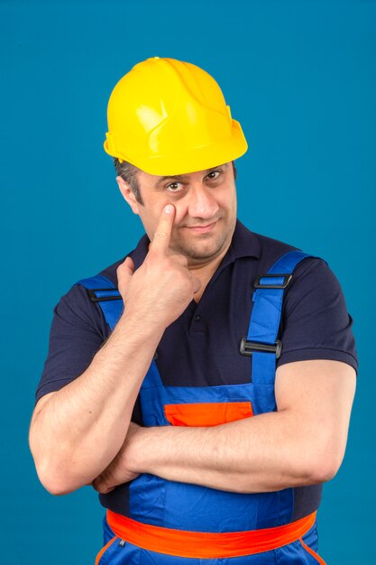 Builder man wearing construction uniform and safety helmet pointing to the eye watching you gesture suspicious expression over isolated blue wall