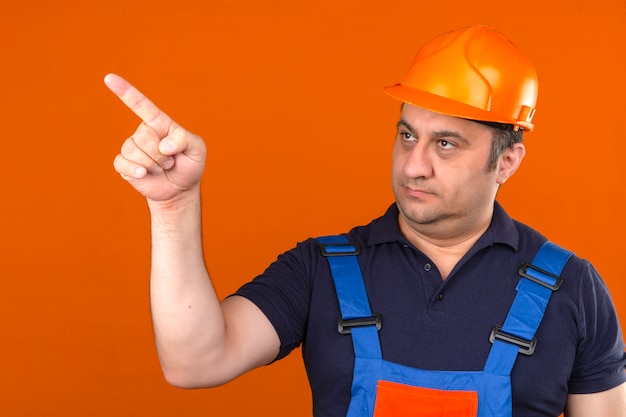 Человек-строитель в строительной форме и защитном шлеме, указывая в сторону, стоит с серьезным лицом над изолированной оранжевой стеной
