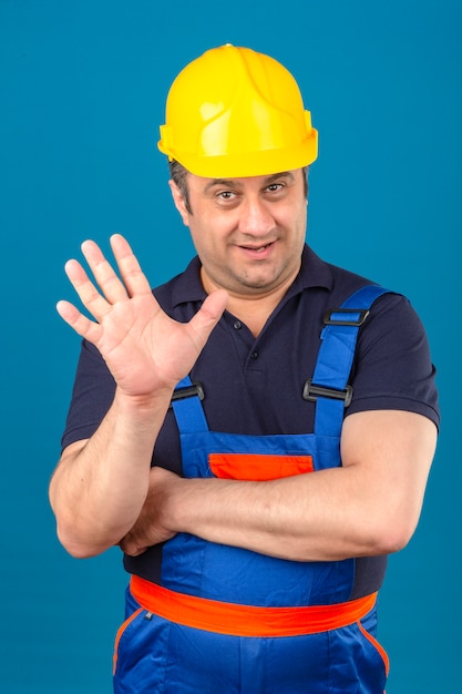 Uniforme da costruzione dell'uomo del costruttore e casco di sicurezza che mostrano e che indicano su con le dita numero cinque mentre sorridendo sicuro e felice sopra la parete blu isolata