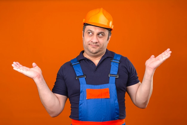 Uomo del costruttore che indossa l'uniforme di costruzione e il casco di sicurezza che scrolla le spalle allargando le mani non capendo cosa è successo espressione confusa e confusa sopra l'arancia isolata