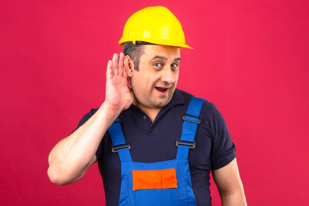 Builder man wearing construction uniform and safety helmet smiling with hand over ear listening an hearing over isolated pink wall