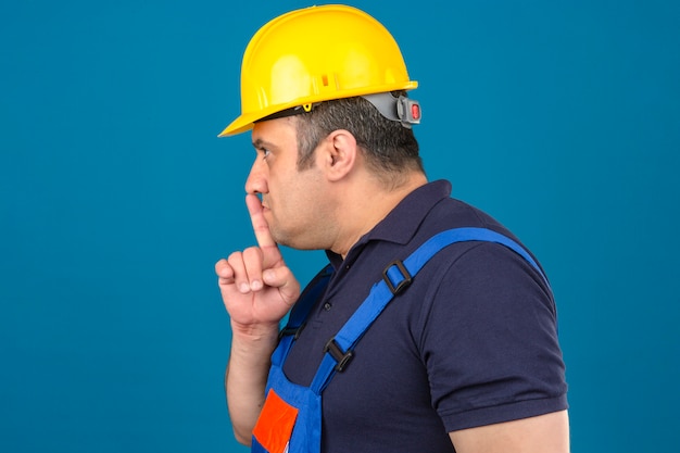 Foto gratuita l'uniforme d'uso della costruzione dell'uomo e il casco di sicurezza che stanno lateralmente che fanno il silenzio firmano sopra la parete blu isolata