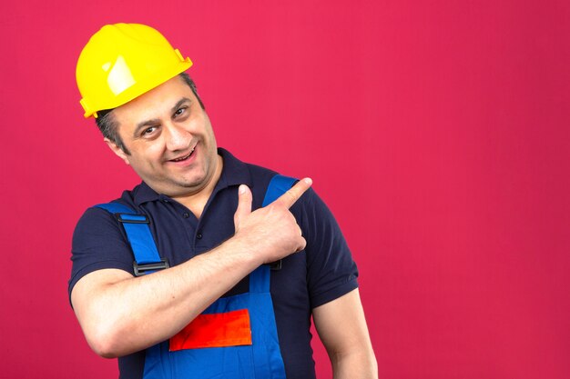 고립 된 분홍색 벽에 손가락으로 측면을 가리키는 얼굴에 큰 미소로 건설 유니폼과 안전 헬멧 서 입고 작성기 남자