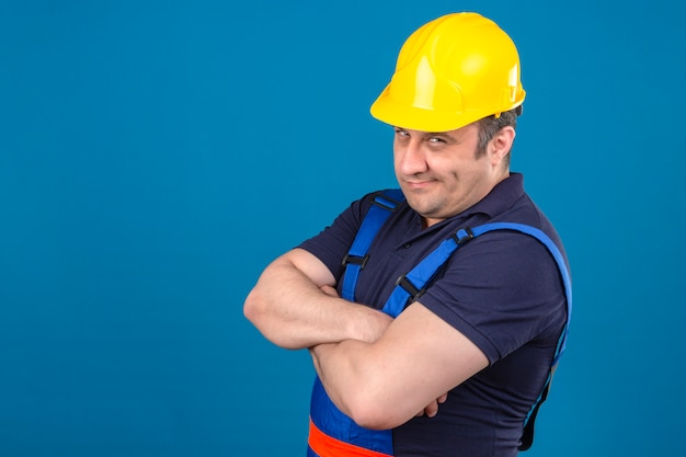 Foto gratuita uniforme d'uso della costruzione dell'uomo e casco di sicurezza del costruttore che stanno con le armi attraversate che ritengono scettico dubbioso sopra la parete blu isolata