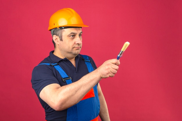 Builder man wearing construction uniform and safety helmet standing with paint brush and pointing with it to the side over isolated pink wall