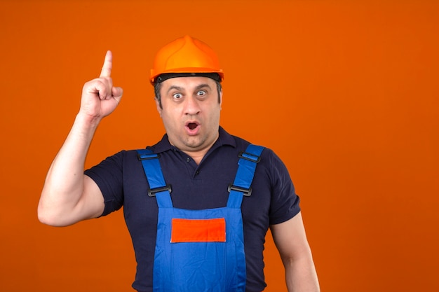 Builder man wearing construction uniform and safety helmet standing with surprised face pointing finger up new idea concept over isolated orange wall