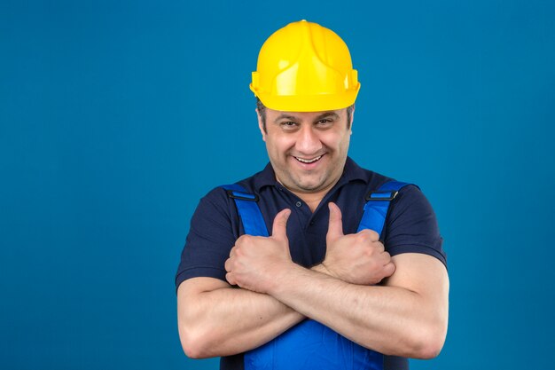 Builder man wearing construction uniform and safety helmet with big smile on face and showing thumbs up over isolated blue wall