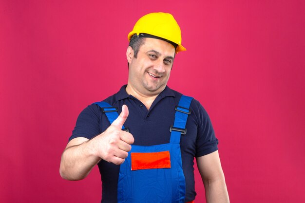 Человек-строитель в строительной форме и защитном шлеме с большой улыбкой на лице и показывает палец вверх над изолированной розовой стеной