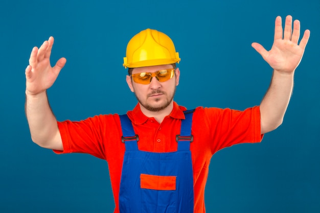 Builder man wearing construction uniform and security helmet arguing have quarrel raising hands looking confused with annoyed bothered face over isolated blue wall