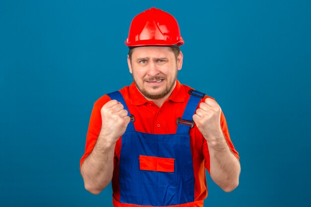 Builder man wearing construction uniform and security helmet displeased clenches fists standing over isolated blue wall