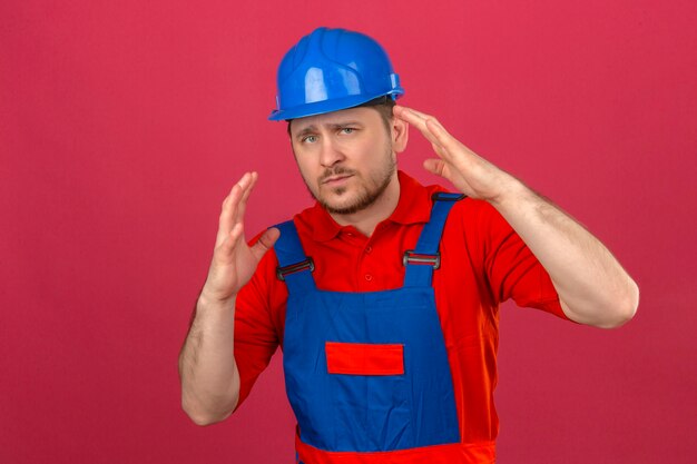 Builder man wearing construction uniform and security helmet displeased and fearful standing with hands raised over isolated pink wall