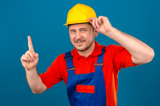 건설 유니폼 및 보안 헬멧을 착용하는 작성기 남자는 고립 된 파란색 벽 위에 서있는 그의 헬멧을 만지고 손가락을 가리키는 아이디어를 얻었습니다.