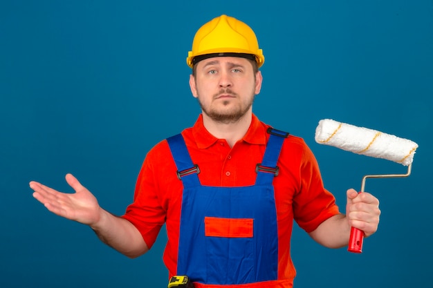 Человек-строитель в строительной форме и защитном шлеме, держащий валик с малярным валиком в руке, невежественное и смущенное выражение с руками и руками вызвал сомнение в концепции над изолированной синей стеной