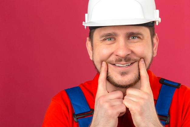 Мужчина-строитель в строительной форме и защитном шлеме держит пальцы на щеках и пытается заставить себя улыбнуться над изолированной розовой стеной