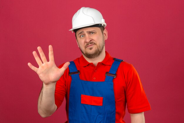 Builder man wearing construction uniform and security helmet showing and pointing up with fingers number five with skeptic expression on face over isolated pink wall