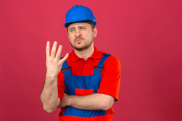 Человек-строитель в строительной форме и защитном шлеме показывает и указывает пальцами номер четыре со скептическим выражением лица над изолированной розовой стеной